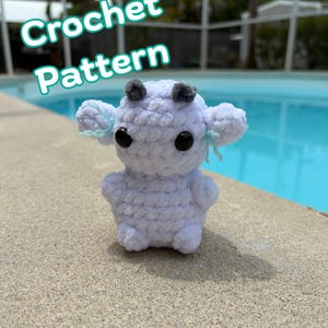 May include: A small, white, plush crochet creature with two dark gray horns, large black eyes, and light blue accents on the ears. The text 'Crochet Pattern' is displayed in white with a teal outline above the plush figure.