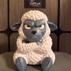 Könnte beinhalten: Eine Schaf-Figur mit einem strukturierten weißen Wollkörper und einem glatten, bläulichen Gesicht mit wütendem Ausdruck, zusammengekniffenen Augenbrauen und verschränkten Armen. Das Schaf sitzt, und ein Schild darüber zeigt CraftLand Studio.