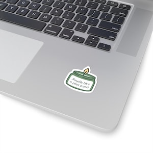 May include: A sticker featuring a green candle in a jar with a lit flame. The text "Smells like a plot twist" is written in white inside the jar. The sticker is placed on a laptop.