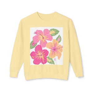 Pode incluir: Camisola amarela clara com um design floral de flores de hibisco rosa e amarelo e folhas verdes sobre um fundo branco. A camisola tem mangas compridas e gola redonda. O texto "Life is Good" está impresso no canto inferior direito.