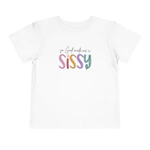 May include: A white short-sleeved t-shirt featuring the black text 'so God made me a' above the colorful, multi-toned word 'SISSY' in a stylized font, with a small flower icon replacing the dot on the letter 'i' in the center of the graphic.