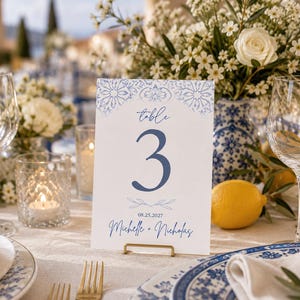 Puede incluir: Una tarjeta de número de mesa rectangular blanca con un patrón floral azul en la parte superior, el texto Table 3, la fecha 08.25.2027 y los nombres Michelle + Nicholas en letra cursiva azul, sostenida por un pequeño soporte de metal dorado.