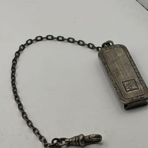 May include: A rectangular silver-toned metal clip with a textured, striped surface and an engraved square emblem. The clip is attached to a thin metal link chain that terminates in a small, rounded swivel clasp, creating a functional accessory.