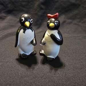 May include: Two ceramic penguin figurines, one labeled Willie and the other labeled Tillie. Both have black and white bodies with yellow beaks. The penguin on the left wears a black bowtie, while the penguin on the right has a red bow and a white beaded necklace.