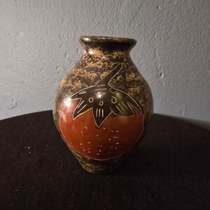 May include: A small, rounded ceramic vase with a narrow neck and a flared rim. The surface features a mottled black and brown finish with a prominent, hand-etched graphic of a red strawberry with green leaves centered on the front of the vessel.