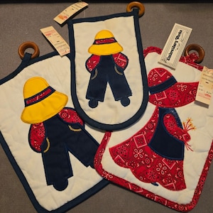 May include: Three quilted fabric potholders with wooden hanging rings. Two feature a figure in a yellow hat and navy blue overalls with red bandana-print sleeves. One features a figure in a red bonnet and dress with red bandana-print accents.
