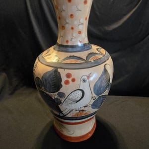 Puede incluir: Un jarrón de cerámica con base color crema que presenta diseños pintados a mano de un pájaro blanco, hojas azul oscuro y puntos naranjas. El cuello tiene un patrón de puntos naranjas y líneas negras. La palabra Mexico está escrita en la base.