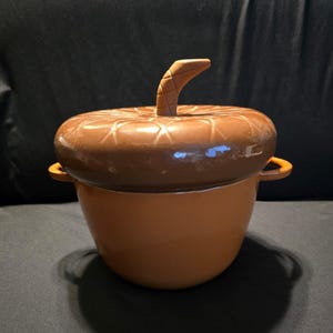 May include: A brown ceramic lidded jar shaped like an acorn. The lid features a textured stem handle and a cross-hatched pattern on the top. The base is a smooth, rounded pot with two small side handles, finished in a glossy, solid brown glaze.