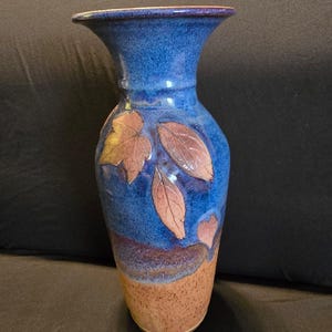 May include: A tall ceramic vase with a flared rim, featuring a vibrant blue glaze on the upper portion and a speckled tan base. The body is decorated with embossed leaf patterns in a muted brown tone, creating a textured contrast against the blue.