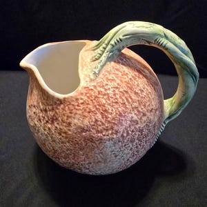 May include: A ceramic pitcher with a round, textured body in shades of mottled brown and tan. The interior is smooth and white. It features a prominent, sculpted handle shaped like a green, gnarled branch or vine that attaches to the top rim.