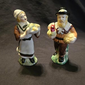 May include: Two ceramic figurines depicting a pair of figures in historical attire. The left figure wears a white bonnet, brown dress, and white apron, holding a bowl. The right figure wears a black hat, brown shirt, and holds a turkey and pumpkin.