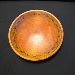 May include: A round wooden bowl with a smooth, light brown finish. The interior rim features a decorative painted border consisting of a wavy yellow line, a thin green line, and small dark rectangular markings spaced evenly around the edge.