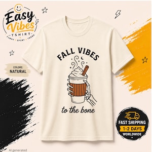 Può includere: Una t-shirt color naturale con la grafica di una mano di scheletro che regge una tazza di caffè con panna montata e una stecca di cannella. Il testo sopra la tazza recita FALL VIBES e il testo sotto la tazza recita to the bone in nero.