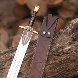 May include: A silver-toned sword with a dark brown handle, a gold-colored crossguard, and a shield-shaped emblem featuring a trident symbol on the blade. The sword is accompanied by a dark brown leather sheath with visible stitching and metal rivets.