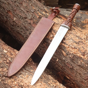 May include: A double-edged steel dagger with a carved dark wood handle featuring a braided pattern and a silver-toned bolster. The dagger is accompanied by a brown leather sheath with visible stitching and a small snap-button strap closure.