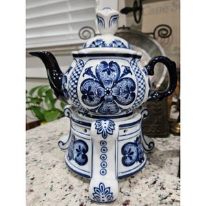 May include: A white ceramic teapot with a matching warmer stand, both decorated with intricate blue floral patterns. The teapot features a dark blue handle and spout, while the base has a circular design with cutouts and ornate blue detailing.