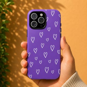 Op de afbeelding: Een paarse smartphonehoes met een herhalend patroon van handgetekende witte hartjes. De hoes heeft een rechthoekige vorm met afgeronde hoeken en een grote ronde uitsparing in de linkerbovenhoek voor de cameralenzen van de telefoon.