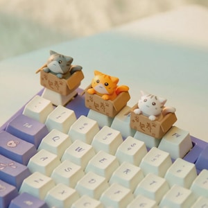 May include: Three miniature cat figurines, one grey, one orange, and one white, each sitting inside a small brown cardboard box with Chinese characters printed on the side, attached as decorative keycaps on a purple and white computer keyboard.