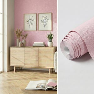 May include: A roll of textured light pink wallpaper with a matte finish shown next to a room scene featuring a light wood sideboard, two framed botanical line art prints on a pink wall, a potted plant, and an open book on a table.
