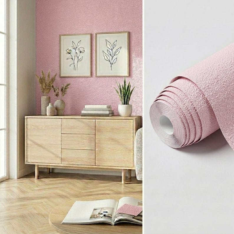 May include: A roll of textured light pink wallpaper with a matte finish shown next to a room scene featuring a light wood sideboard, two framed botanical line art prints on a pink wall, a potted plant, and an open book on a table.