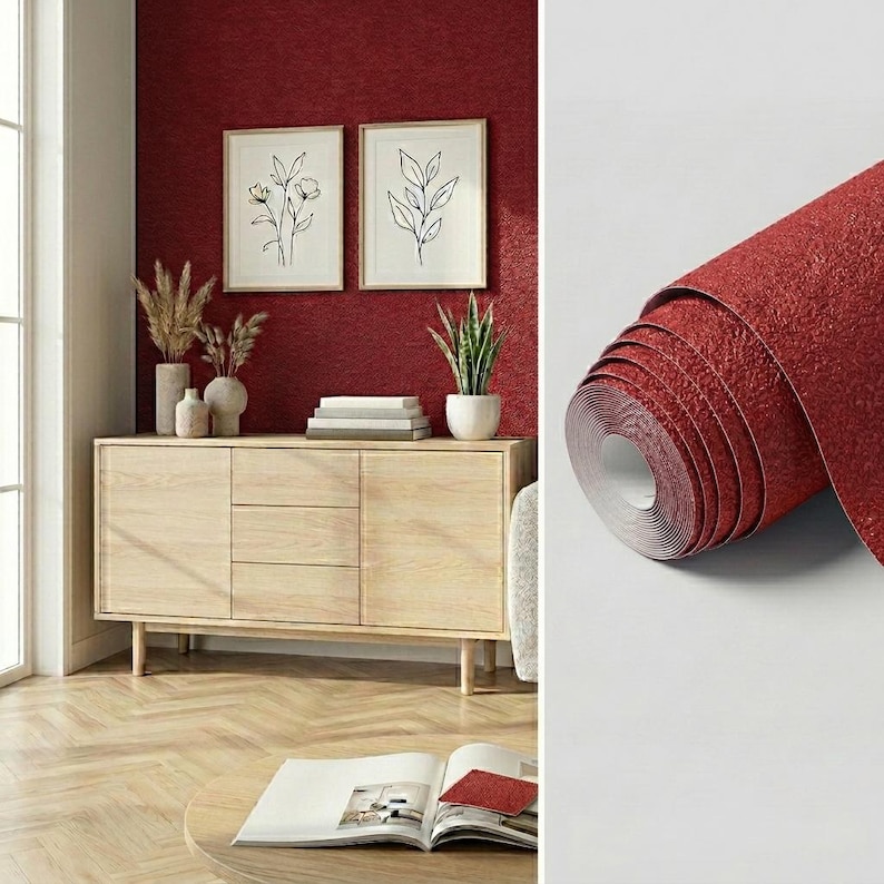 May include: A roll of deep red wallpaper with a textured, granular surface finish. The wallpaper is shown in a room setting featuring a light wood sideboard, two framed botanical line art prints, and a book on a round wooden table.