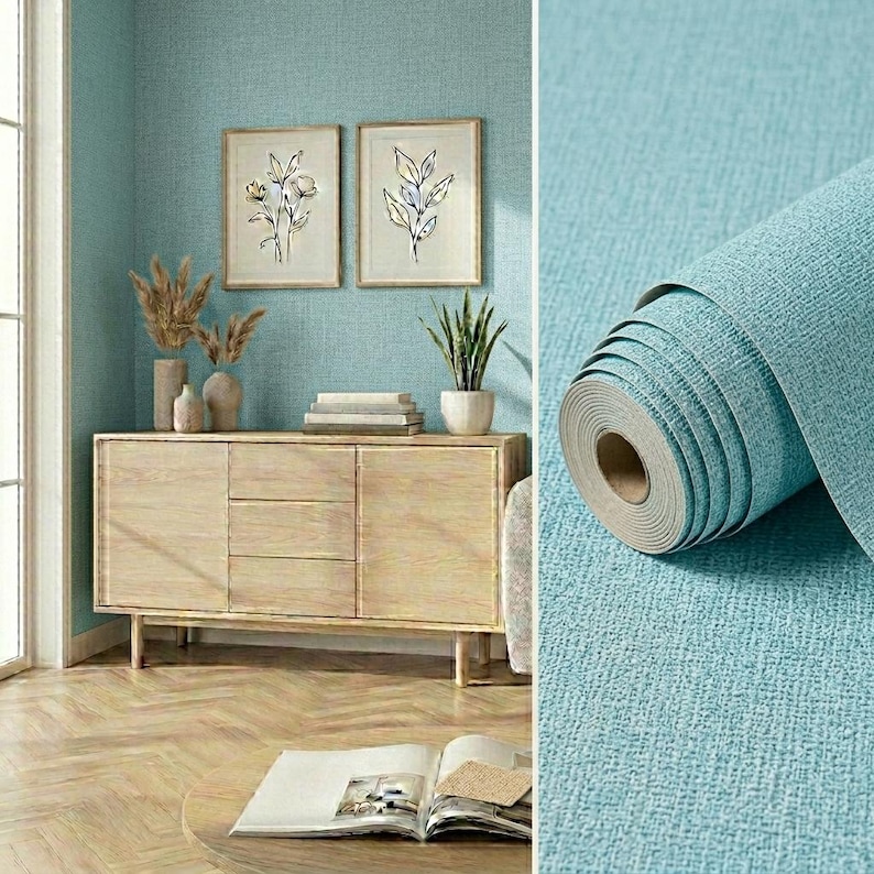 May include: A roll of textured teal wallpaper with a woven fabric appearance. The wallpaper is shown in a room setting featuring a light wood sideboard, two framed botanical line art prints, a potted plant, and a book on a wooden table.