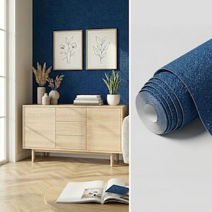 May include: A roll of dark blue wallpaper with a textured surface shown next to a room scene featuring a light wood sideboard, two framed botanical line art prints, a potted plant, and a vase with dried grasses on a blue wall.
