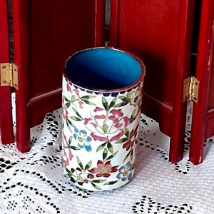May include: A cylindrical metal vessel featuring a white exterior decorated with a colorful floral pattern of pink, red, and blue flowers with green leaves. The interior of the vessel is a solid, vibrant turquoise blue color with a metallic rim.