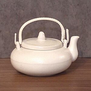May include: A white ceramic teapot with a rounded body featuring a subtle dimpled texture. It includes a matching lid with a small knob handle and a curved spout. A white woven-style handle is attached to two upright metal brackets.