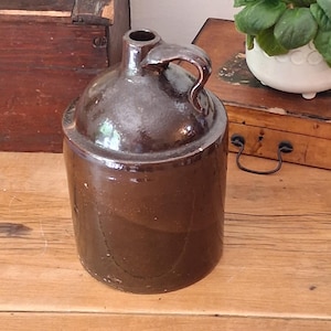 May include: A dark brown glazed ceramic jug with a cylindrical body, a rounded shoulder, and a single handle attached to the neck. The surface has a smooth, reflective finish with subtle variations in the deep brown color throughout the piece.