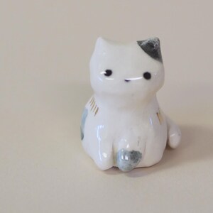 May include: A small white ceramic cat figurine with a glossy finish. The cat features simple black dot eyes, a small grey patch on its head, and subtle grey and gold painted accents on its body. The cat is depicted in a seated position.