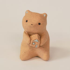 May include: A small, light brown ceramic bear figurine with a speckled texture. The bear has two tiny black eyes and holds a miniature house in its paws. The house is light brown with a pale blue roof and a small blue square window on its front.
