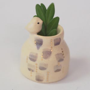 May include: A small, cream-colored ceramic planter featuring a sculpted bird on the rim. The exterior is decorated with a pattern of muted purple brushstrokes and vertical lines of small gold dots. A green succulent plant is placed inside the vessel.