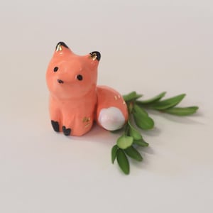 May include: A small, hand-painted ceramic fox figurine with a bright orange body, black-tipped ears, black paws, and a white-tipped tail. The fox has gold-painted accents on its ears and side, and it is positioned next to a small green sprig.
