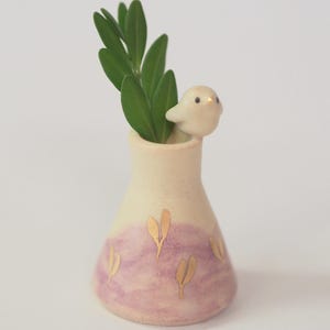 May include: A small ceramic bud vase with a tapered neck and a wide base. The lower portion features a soft lavender wash with gold leaf leaf-shaped accents. A small, sculpted white bird sits on the rim, and green leaves are placed inside.