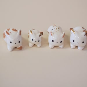 May include: Four small, white ceramic bunny figurines with brown spots and gold accents. Each bunny has black eyes and a cute, rounded shape. The bunnies are arranged in a row on a light beige surface.