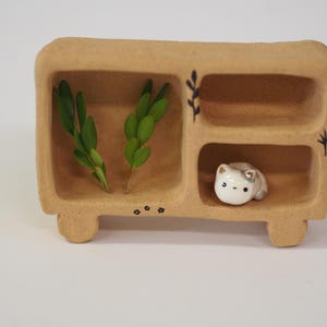 May include: A tan, textured ceramic shelf unit with three open compartments. The left compartment contains two green leafy sprigs. The bottom right compartment holds a small white ceramic cat figurine with gold accents. Black line art details decorate the exterior.