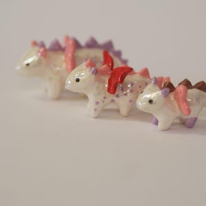 May include: Three small, pearlescent white dragon figurines arranged in a row. Each creature features a glossy finish, small wings, and a row of spikes along its back. The dragons are accented with various colors including purple, red, and brown.
