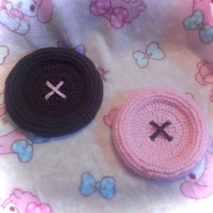May include: Two circular, crocheted button-shaped coasters. One is dark brown with a light pink stitched cross in the center, and the other is light pink with a dark brown stitched cross in the center. Both have a raised, textured edge.