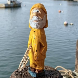 May include: A hand-carved wooden figurine of a person wearing a yellow hooded raincoat and matching trousers. The figure has a white beard, painted facial features, and holds a small piece of tan rope in one hand while standing on a blue base.