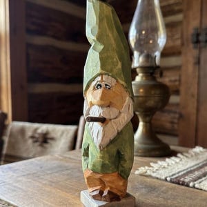 May include: A hand-carved wooden gnome figurine featuring a tall green pointed hat, a white beard, and a brown cigar in its mouth. The gnome wears a green shirt and orange trousers, standing on a square wooden base with visible chisel marks.