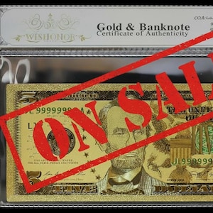 May include: A gold-colored five-dollar banknote replica featuring Abraham Lincoln, enclosed in a clear protective sleeve with a certificate of authenticity at the top. The text ON SALE is stamped in red across the center of the gold banknote.