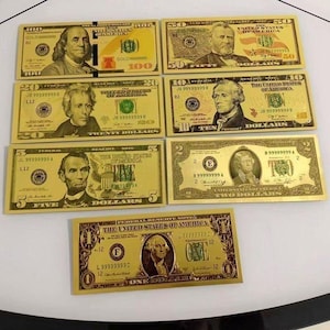 May include: A collection of seven gold-colored novelty banknotes featuring United States currency denominations including one, two, five, ten, twenty, fifty, and one hundred dollar bills, each displaying portraits of historical figures and official text.