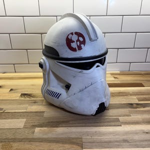 May include: A white clone trooper helmet featuring a weathered finish, a dark visor, and a circular red emblem on the forehead. The helmet includes a central ridge, black accents along the brow, and blue striped detailing on the side cheek area.