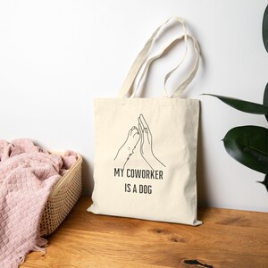 May include: A cream-colored canvas tote bag featuring a black line art illustration of a human hand high-fiving a dog paw. Below the graphic, the text reads "MY COWORKER IS A DOG" in a simple, bold, sans-serif black font. The bag has two handles.