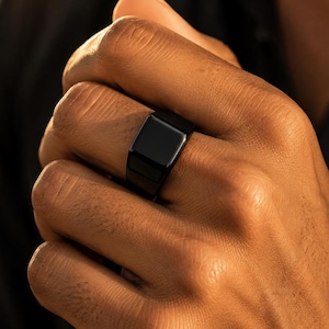 Könnte beinhalten: Ein massiver schwarzer Siegelring mit einer glatten, polierten quadratischen Oberfläche und einer breiten, gleichmäßigen Ringschiene. Der Ring hat ein schlichtes, monochromes Finish mit einer reflektierenden Oberfläche, die das Licht einfängt.