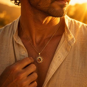 Peut inclure: Un collier en métal doré doté d'un pendentif en forme de soleil avec une pierre précieuse circulaire brune au centre. Le pendentif est suspendu à une fine chaîne dorée, reposant sur une chemise boutonnée beige clair à la texture visible.