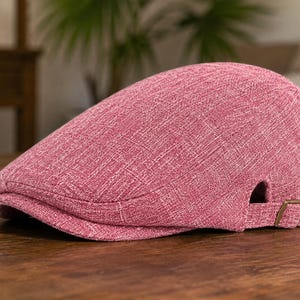 May include: A solid pink flat cap with a textured fabric finish. The cap features a rounded crown, a short stiff brim at the front, and an adjustable strap on the side with a small metallic buckle for securing the fit around the head.