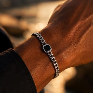 Peut inclure: Un bracelet à maillons gourmette de couleur argentée doté d'un élément central carré avec une incrustation noire lisse. Les maillons métalliques sont polis et interconnectés, formant une bande continue qui entoure le poignet avec un design uniforme.