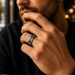 Könnte beinhalten: Ein silberner Ring mit Kettenmuster. Der Ring hat ein rotierendes Mittelband und wird am Finger getragen. Der Ring ist aus Metall und hat eine polierte Oberfläche. Der Ring ist ein Schmuckstück.